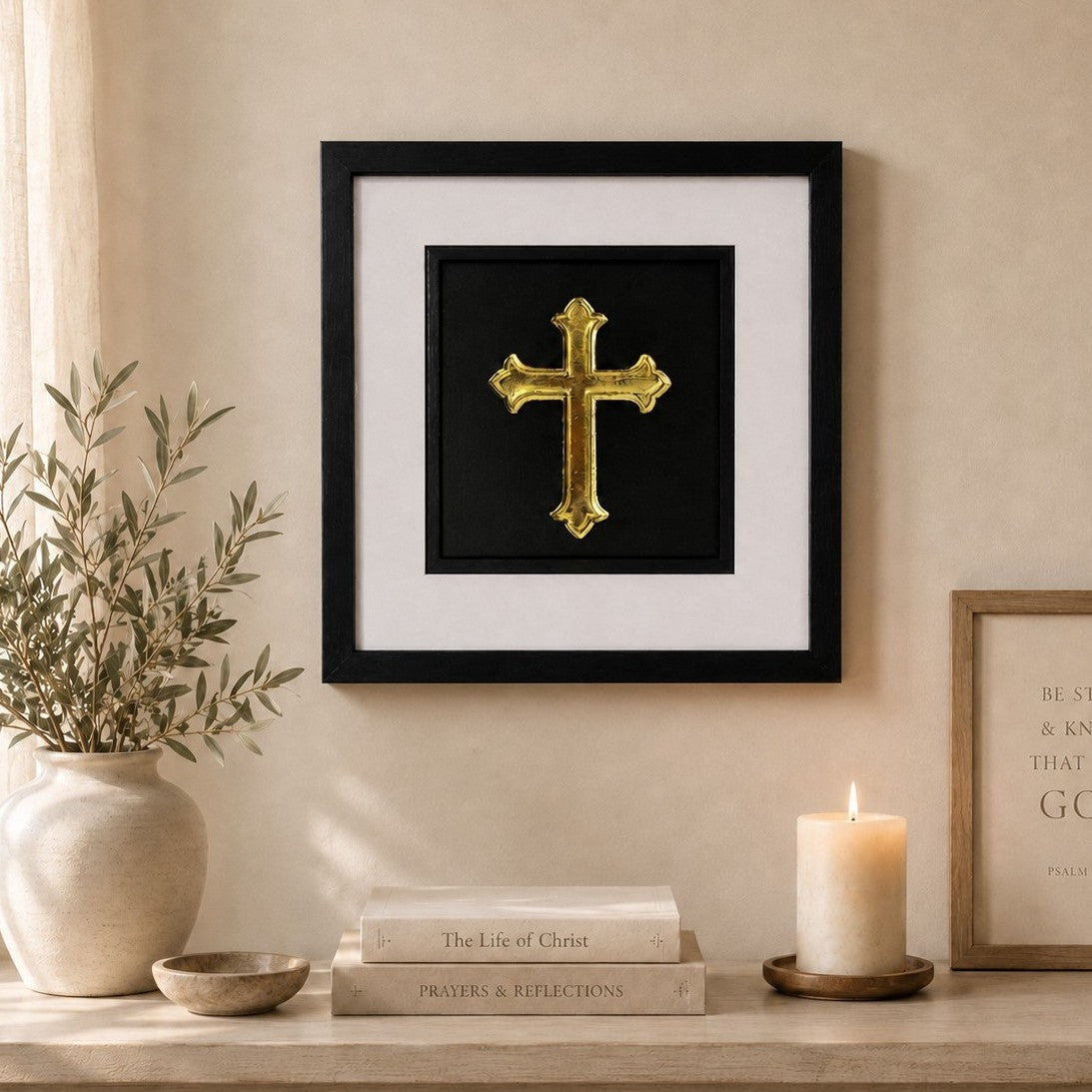 Framed Christian cross wall art finished in gold leaf, styled in a peaceful neutral home setting with candle, olive branches and faith inspired decor.