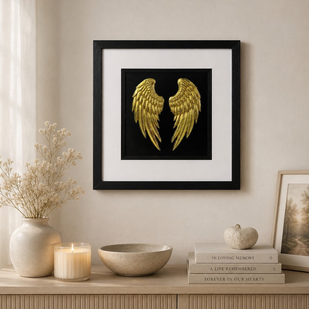 Gold angel wings memorial wall art in a black frame with white mount, styled in a calm neutral home setting with candle, flowers and remembrance decor.