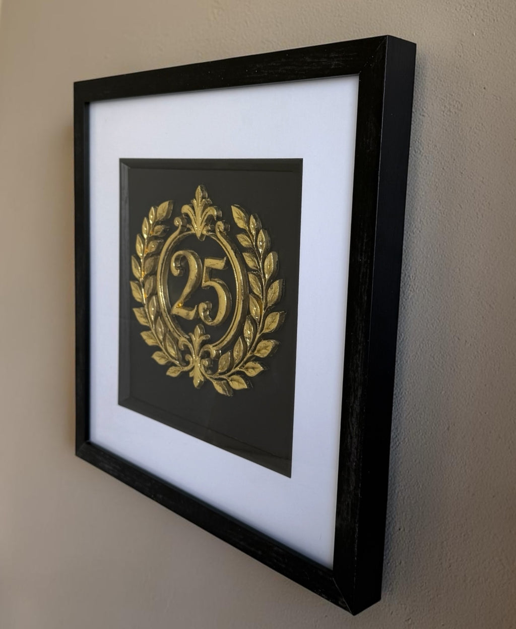 Gold framed 25th anniversary laurel wreath artwork hanging on a wall, showing the raised number 25, black box frame, white mount, and glass front.