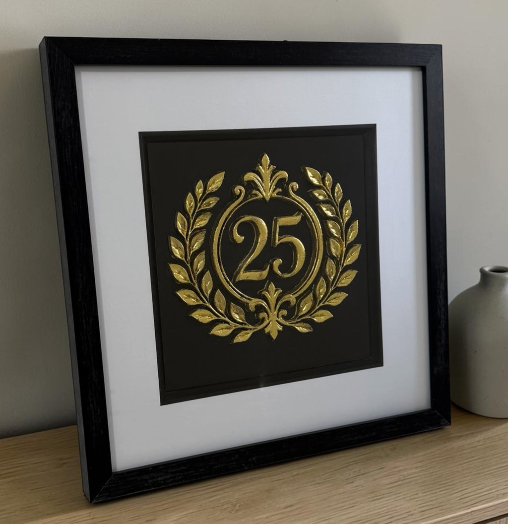Gold 25th anniversary laurel wreath framed artwork leaning on a wooden surface beside a neutral vase, showing frame depth and reflective glazing.