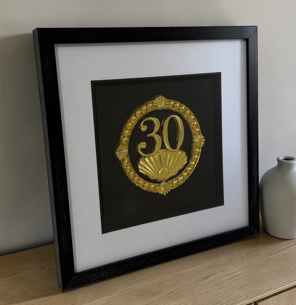 Gold 30th anniversary pearl shell framed artwork leaning on a wooden surface beside a neutral vase, showing frame depth and reflective glazing.