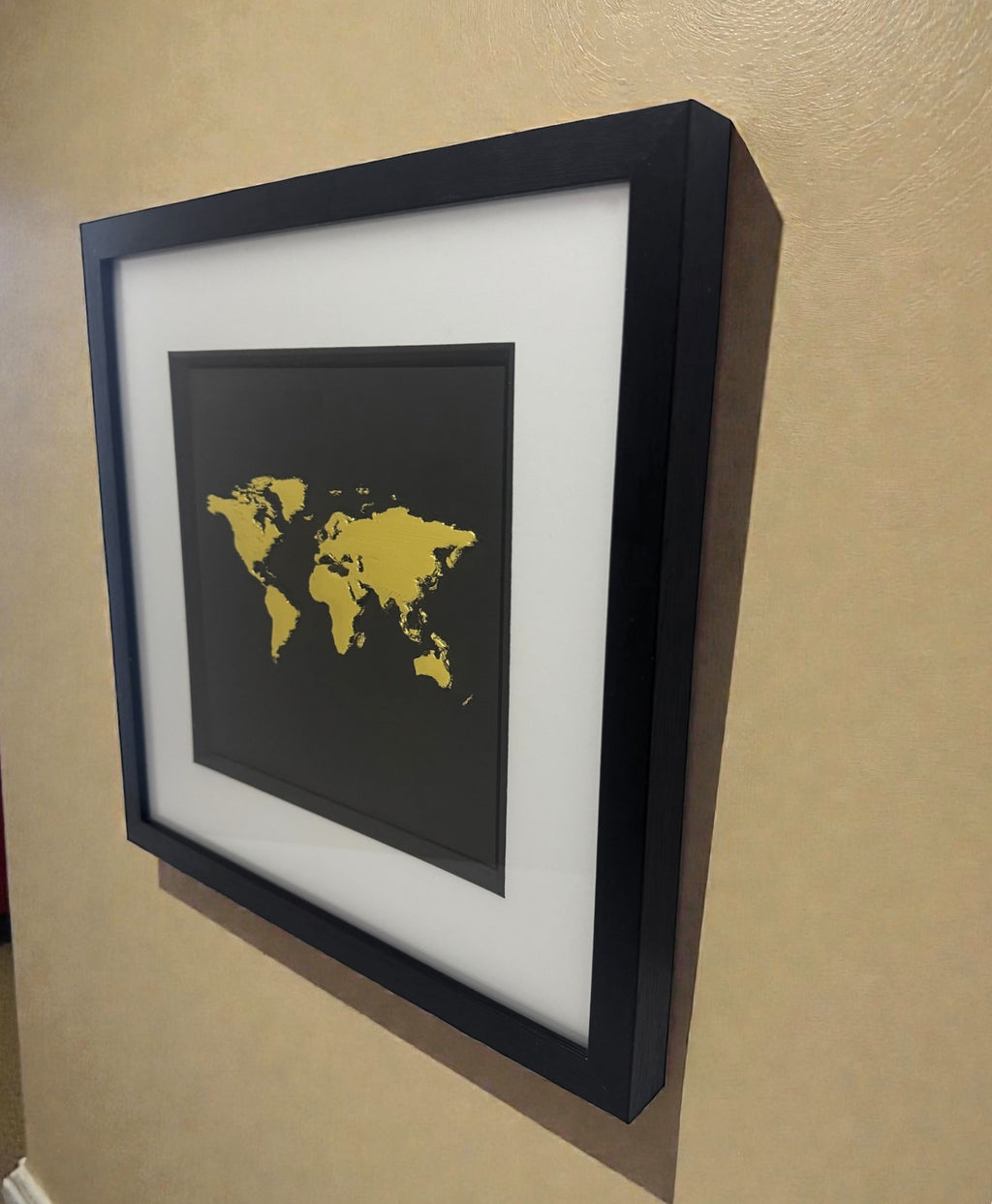 Angled side view of Gold World Map art showing raised 3D relief texture. Hand-gilded metallic leaf sculpture in deep box frame hanging on wall.