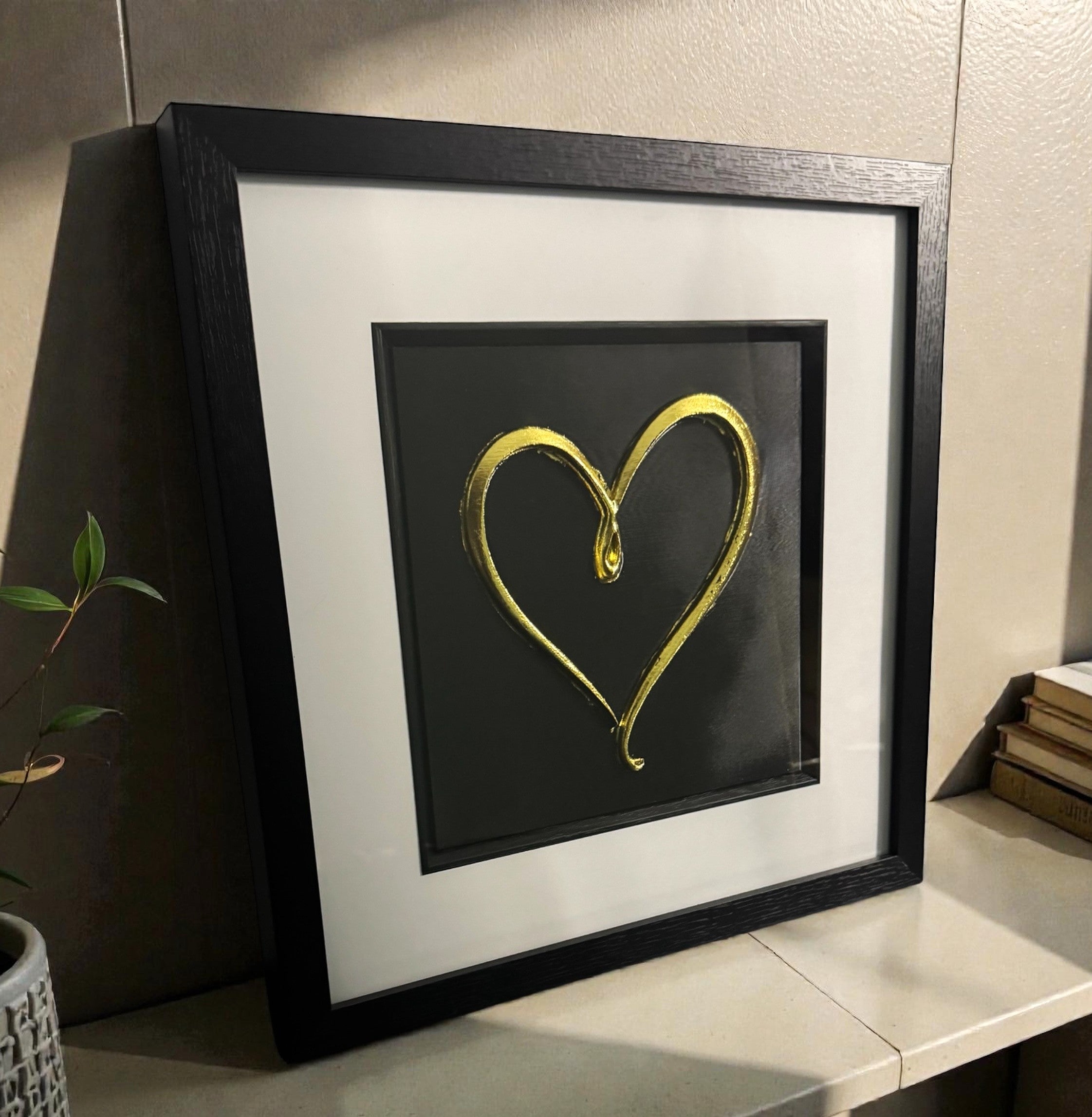 Angled desk view of gold leaf single line heart wall art on a black background in a black wooden frame