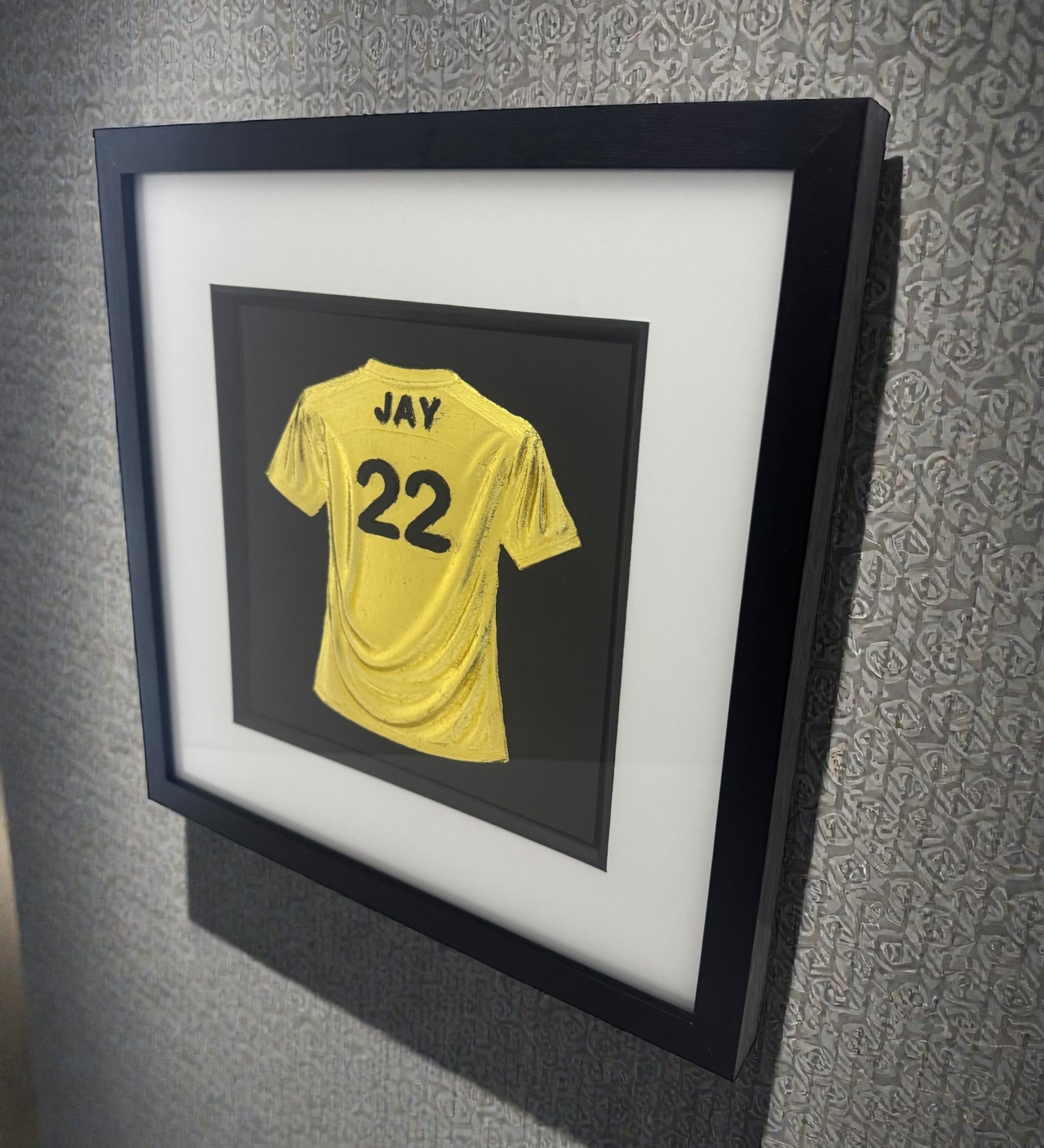 Side view of a framed personalised yellow football shirt 3D wall art with name “JAY” and number “22”, in a black frame with white mount, displayed on a grey wall.