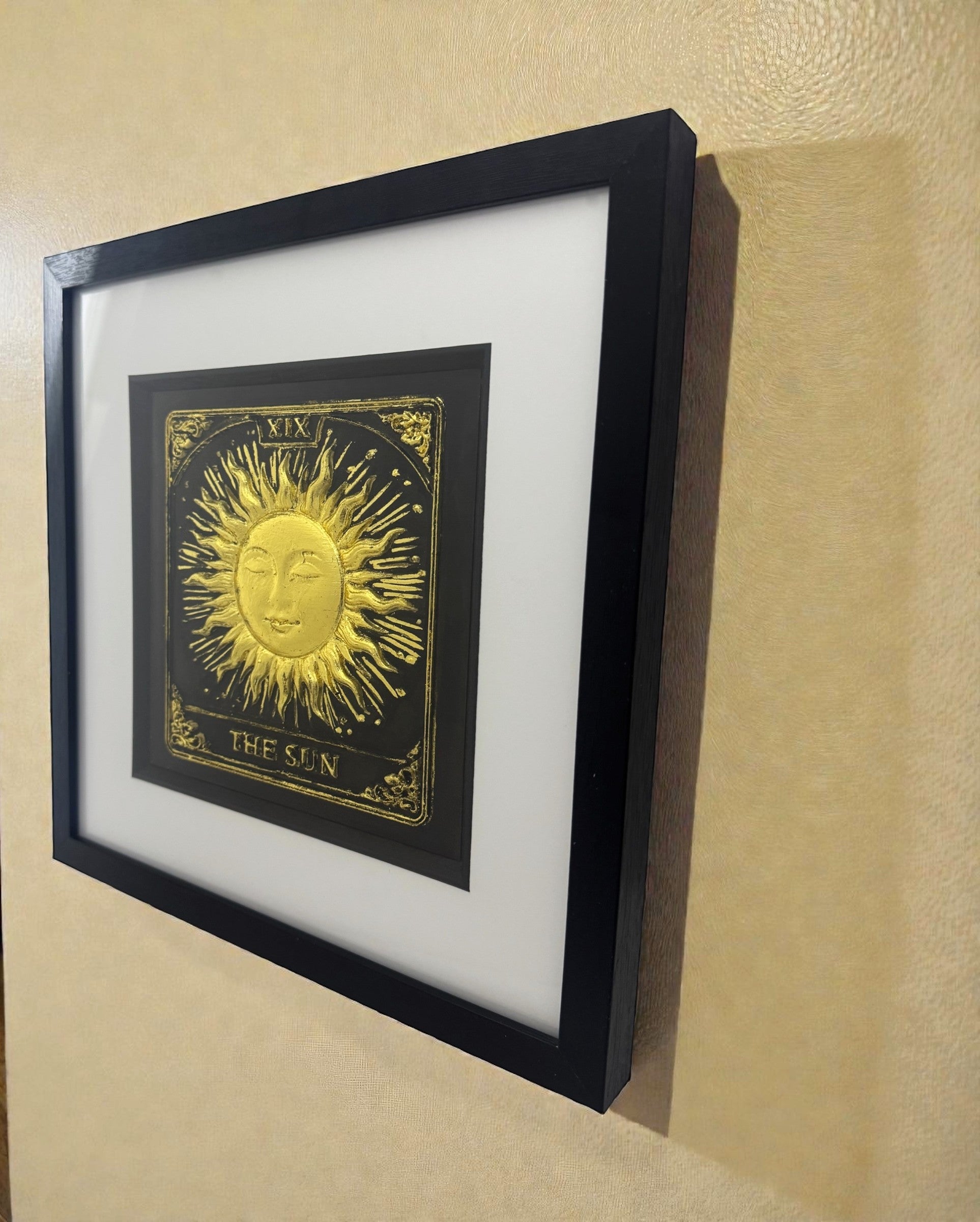 Close-up side view of the 3D relief texture of a gold-leafed Sun Tarot card hanging on a wall, showing the architectural depth.