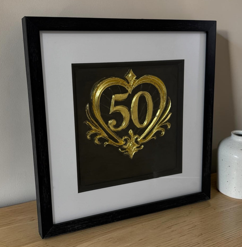 Gold framed 50th anniversary heart artwork leaning on a wooden surface beside a neutral vase, showing the glass front and frame depth.