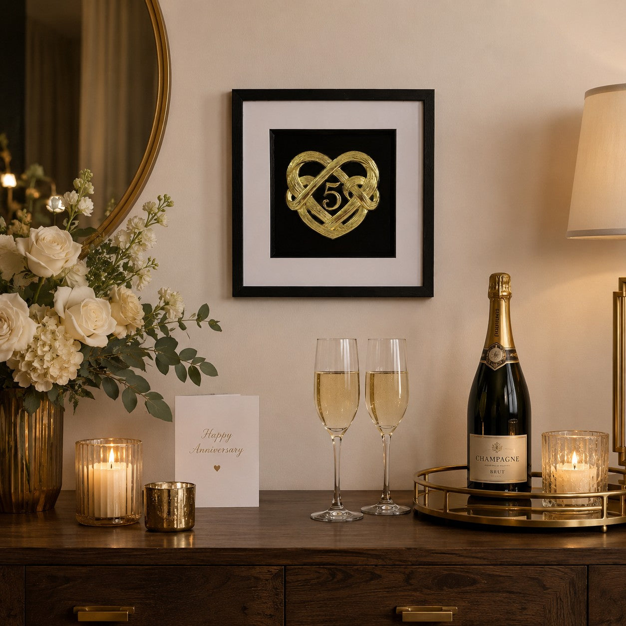 Gold 5th anniversary heart knot framed artwork styled above a wooden sideboard with flowers, candles, champagne glasses, and warm lighting.