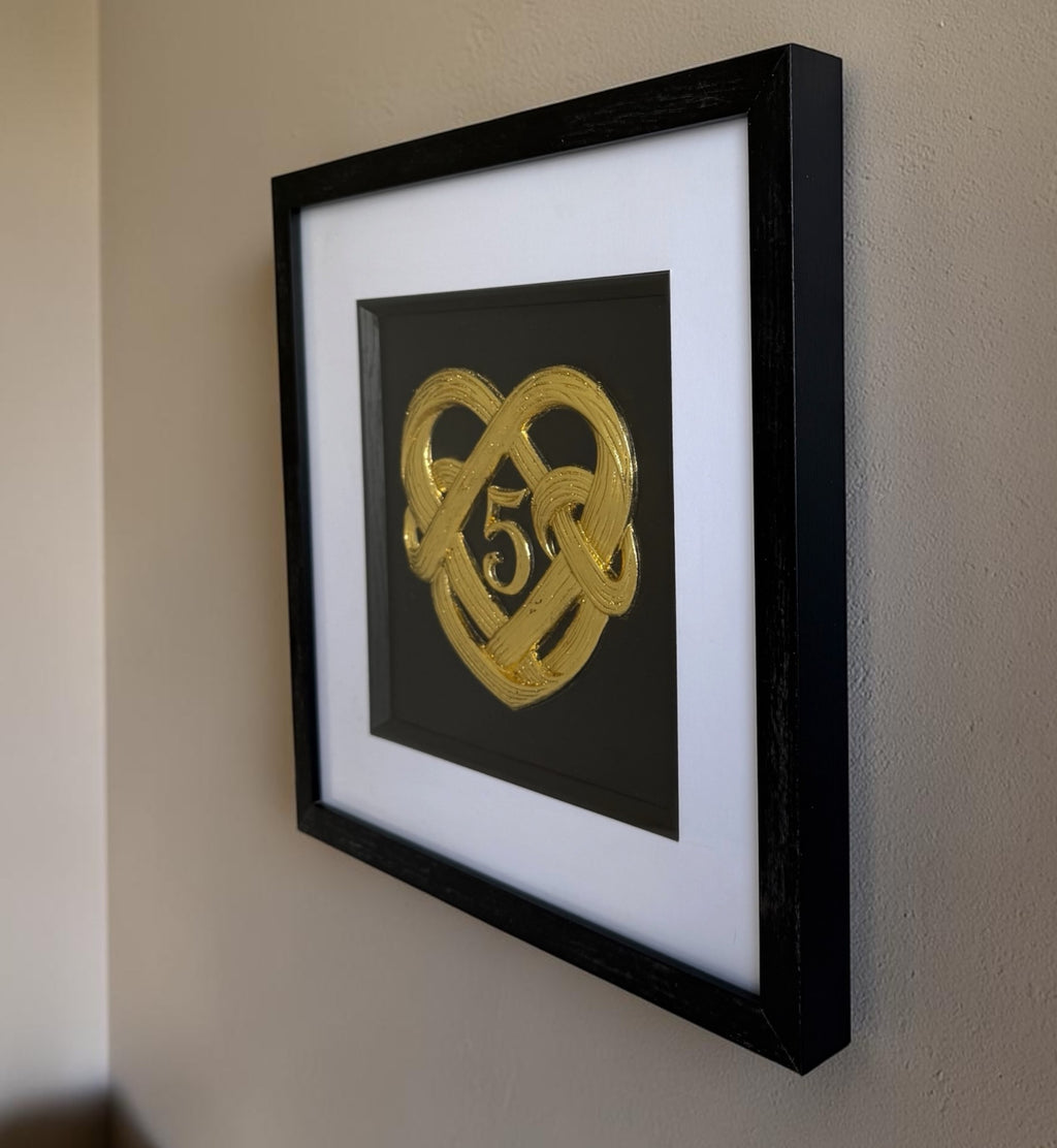 Gold 5th wedding anniversary heart knot artwork hanging on a wall, showing the black box frame depth and white mount from the side.