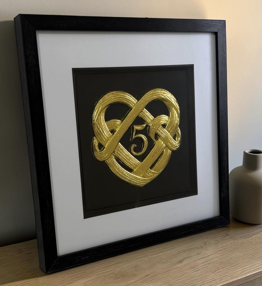 Angled Gold 5th anniversary heart knot framed artwork on a wooden surface showing the glass front, raised detail, and black frame depth.