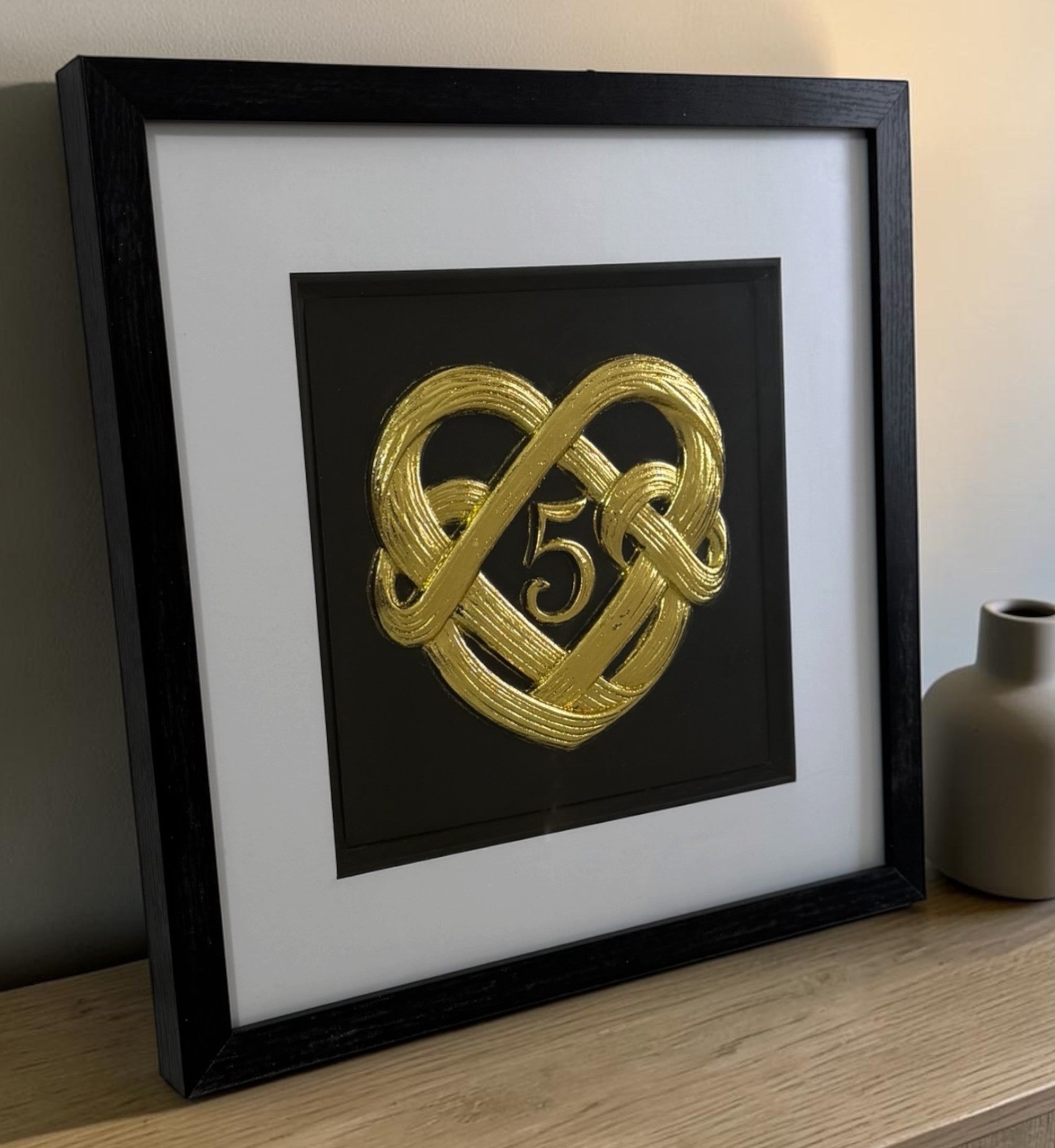 Angled Gold 5th anniversary heart knot framed artwork on a wooden surface showing the glass front, raised detail, and black frame depth.