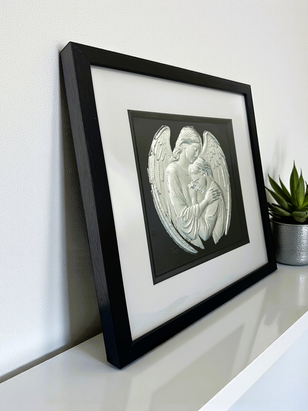 Angled view of angel embrace wall art on a shelf, black frame with white mount and silver leaf finish