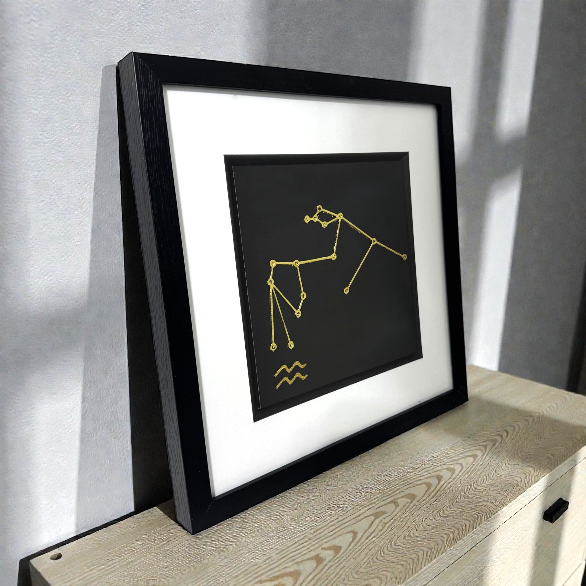 Gold Aquarius constellation wall art shown at an angle on a wooden surface, framed with white mount and raised zodiac star design