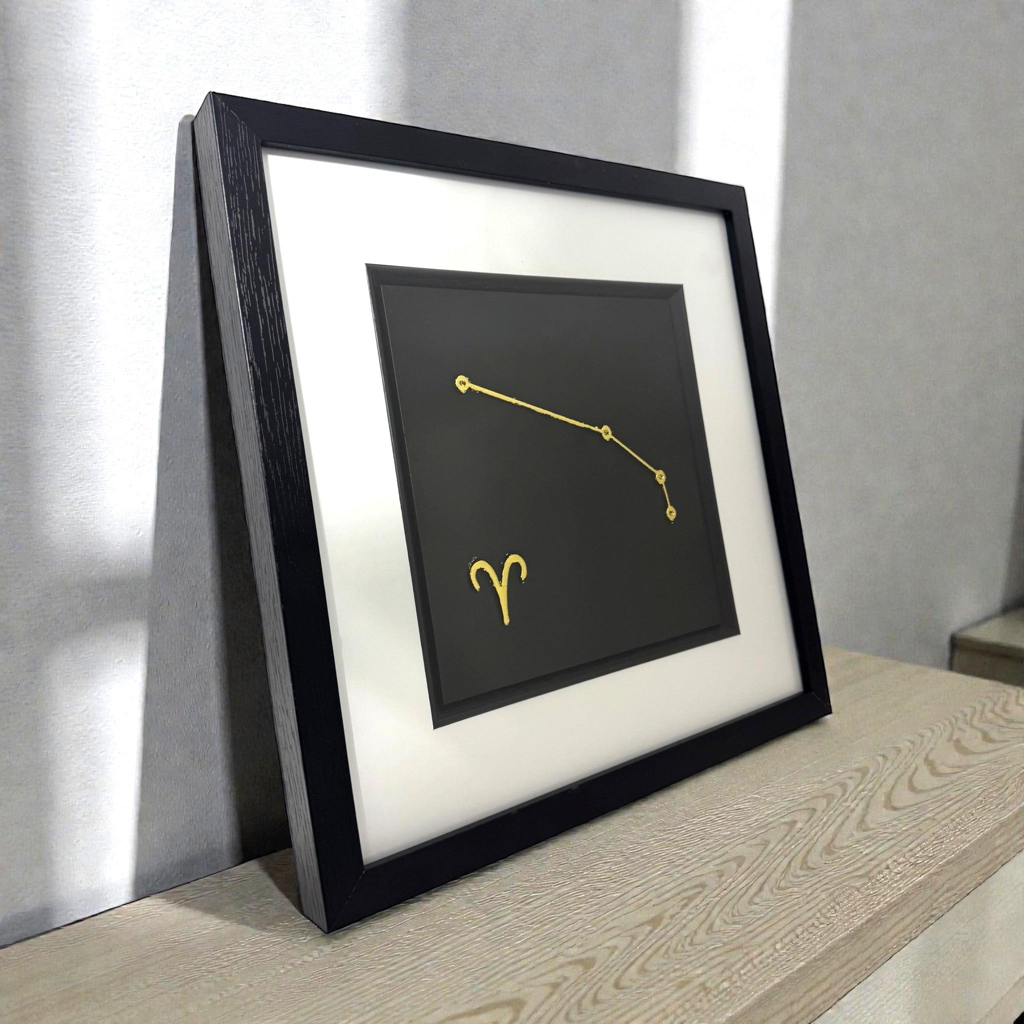 Framed Aries constellation wall art in gold leaf with zodiac symbol on a black background, displayed on a shelf in a modern interior