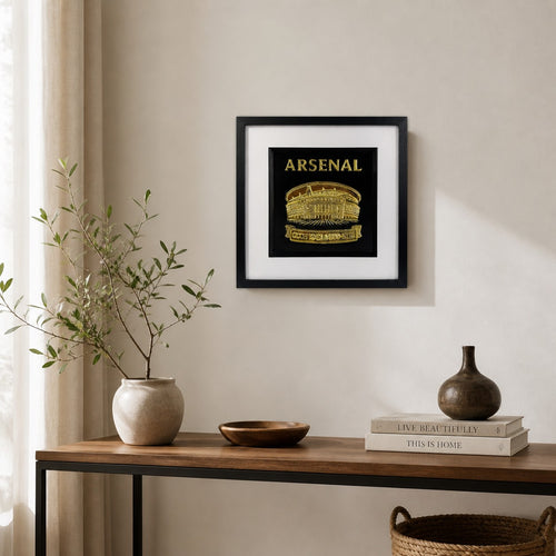 Gold Arsenal Emirates Stadium wall art in a black square frame with white mount, displayed on a neutral wall above a wooden console table.