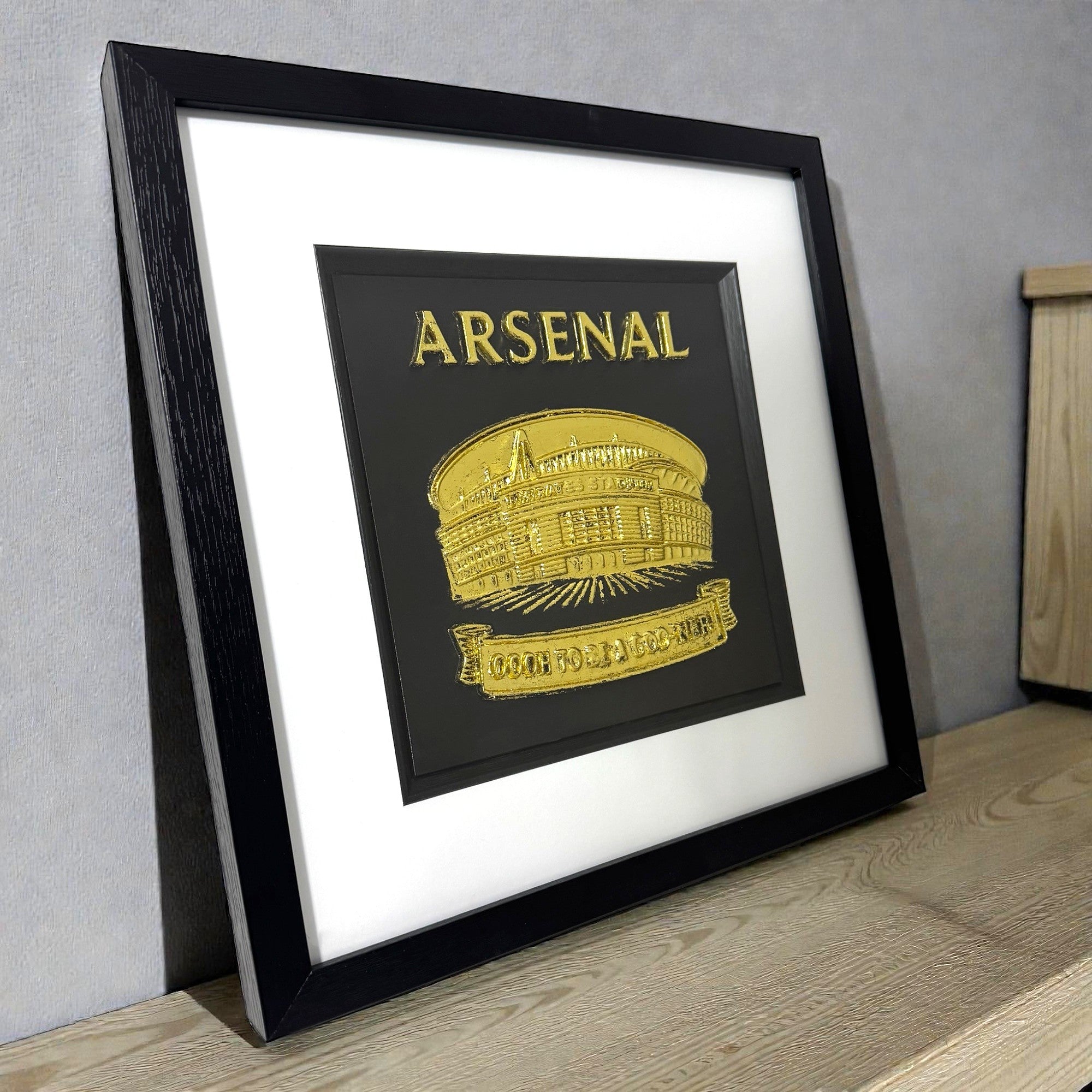 Gold leaf Arsenal stadium wall art displayed on desk in black frame