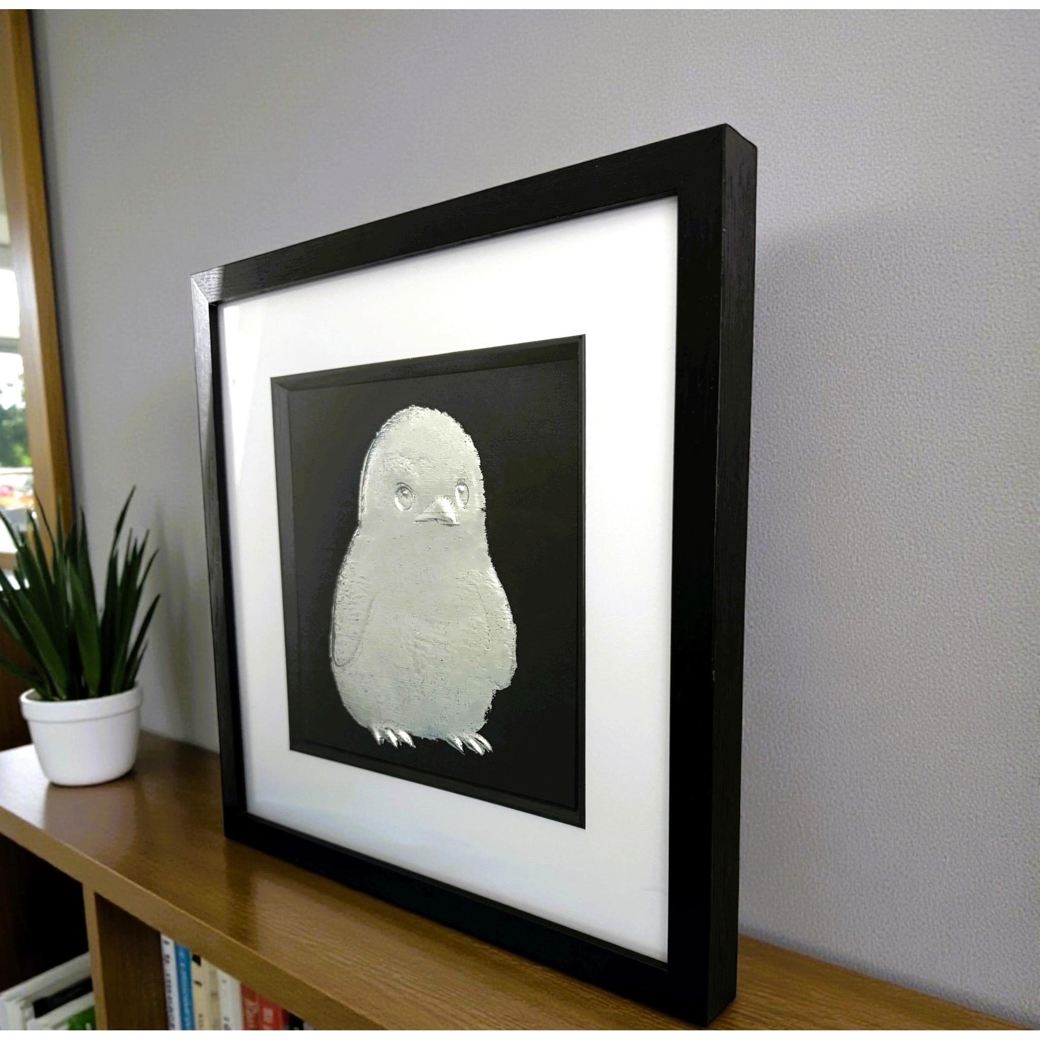 Silver baby penguin wall art framed in black with white mount on a shelf