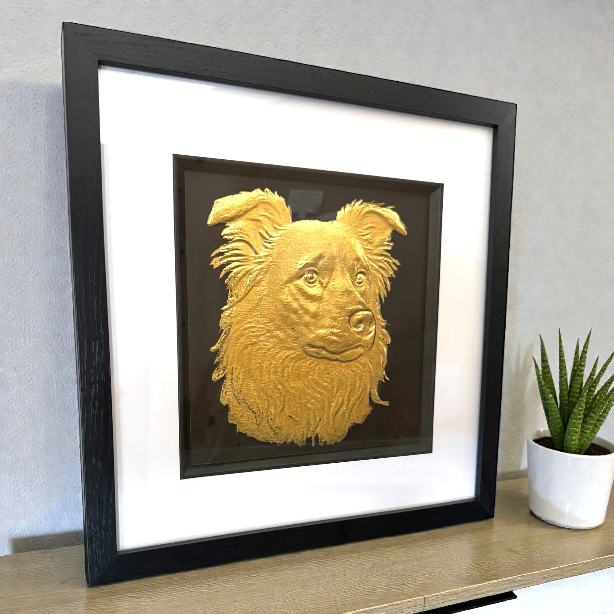 Framed border collie wall art in gold leaf relief on a black background with white mount and black frame, displayed on a shelf in a modern interior