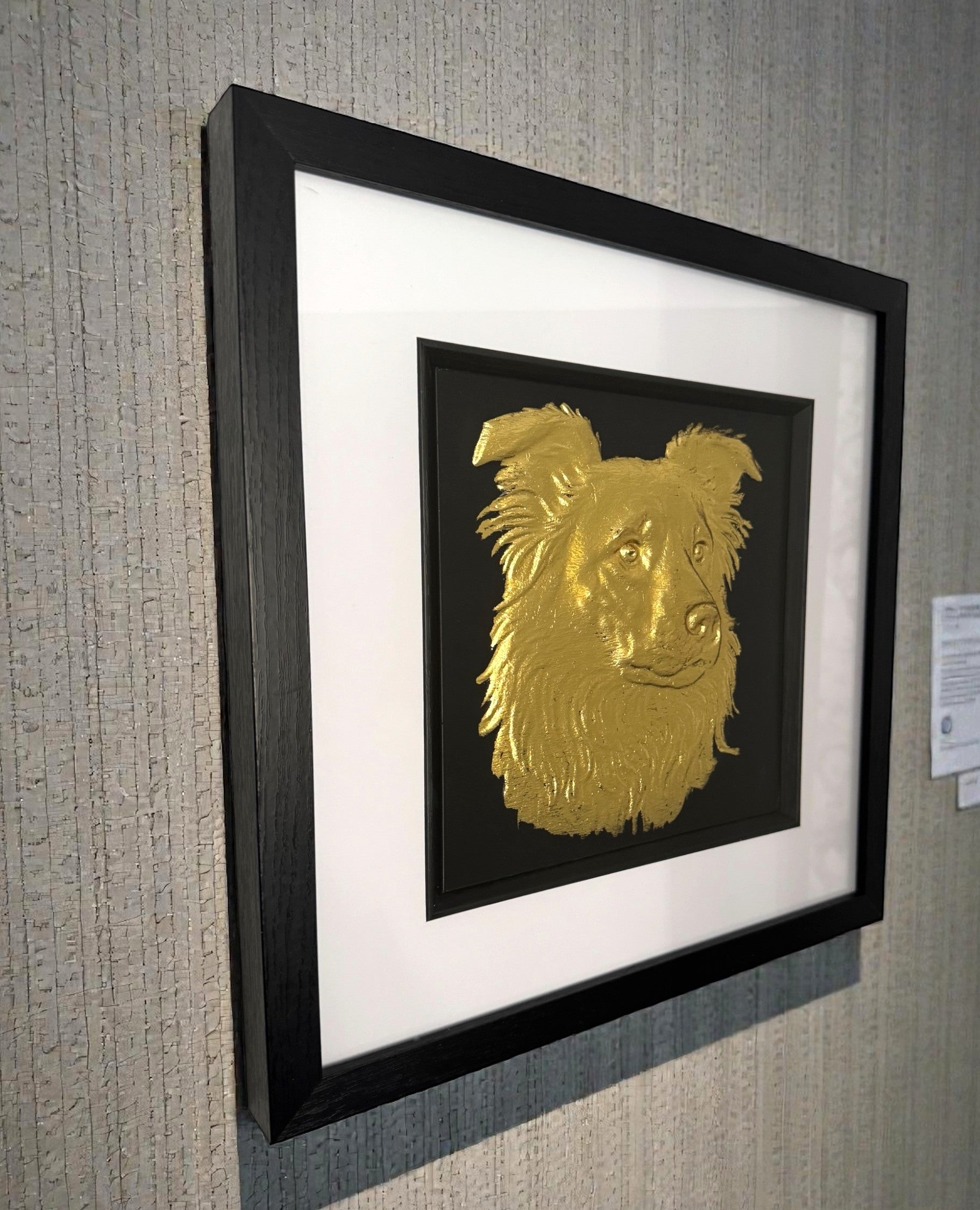 Framed border collie wall art in gold leaf relief mounted on a wall, shown at an angle highlighting the depth and texture of the artwork