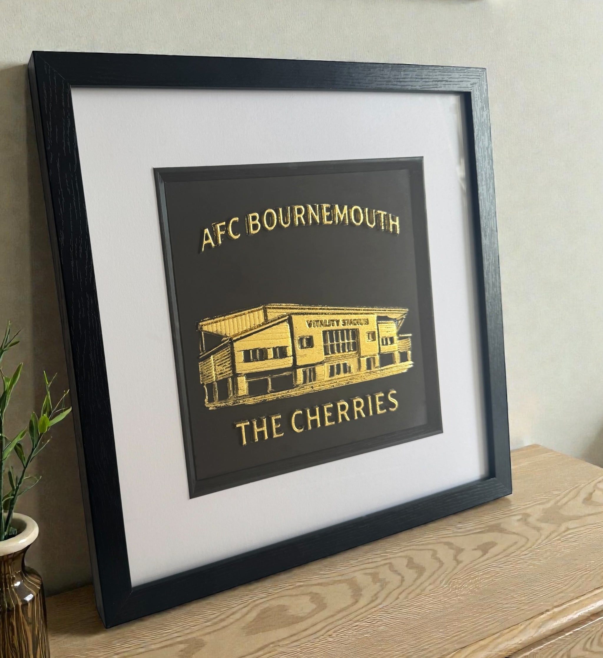 Angled view of framed AFC Bournemouth stadium relief artwork in Gold Leaf with black box frame and white mount, styled on a wooden sideboard