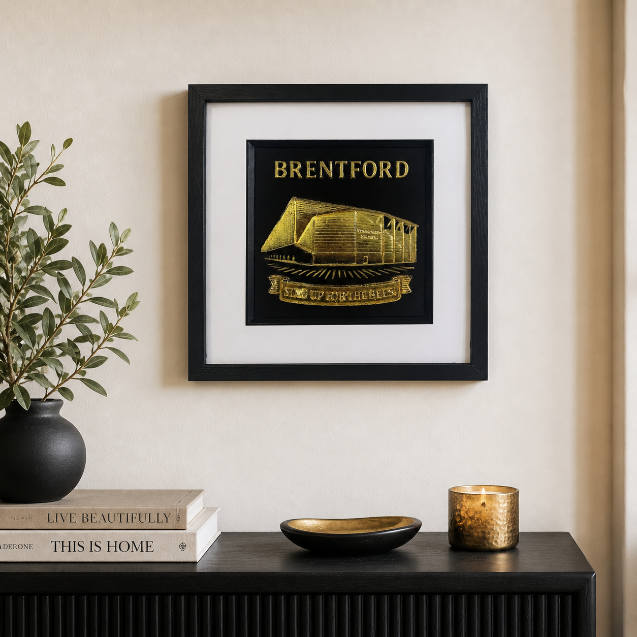 Lifestyle photo of Brentford FC gold wall art displayed on a neutral wall above a black sideboard with plant and candle.