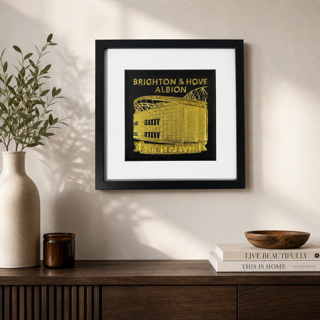 Lifestyle photo of Brighton & Hove Albion gold wall art displayed on a neutral wall above a wooden sideboard.