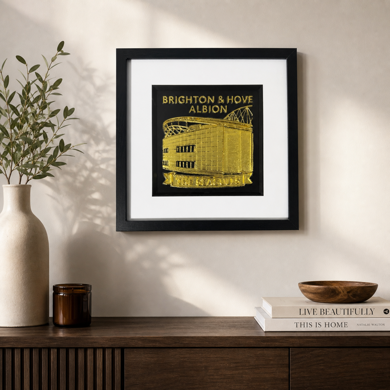 Lifestyle photo of Brighton & Hove Albion gold wall art displayed on a neutral wall above a wooden sideboard.