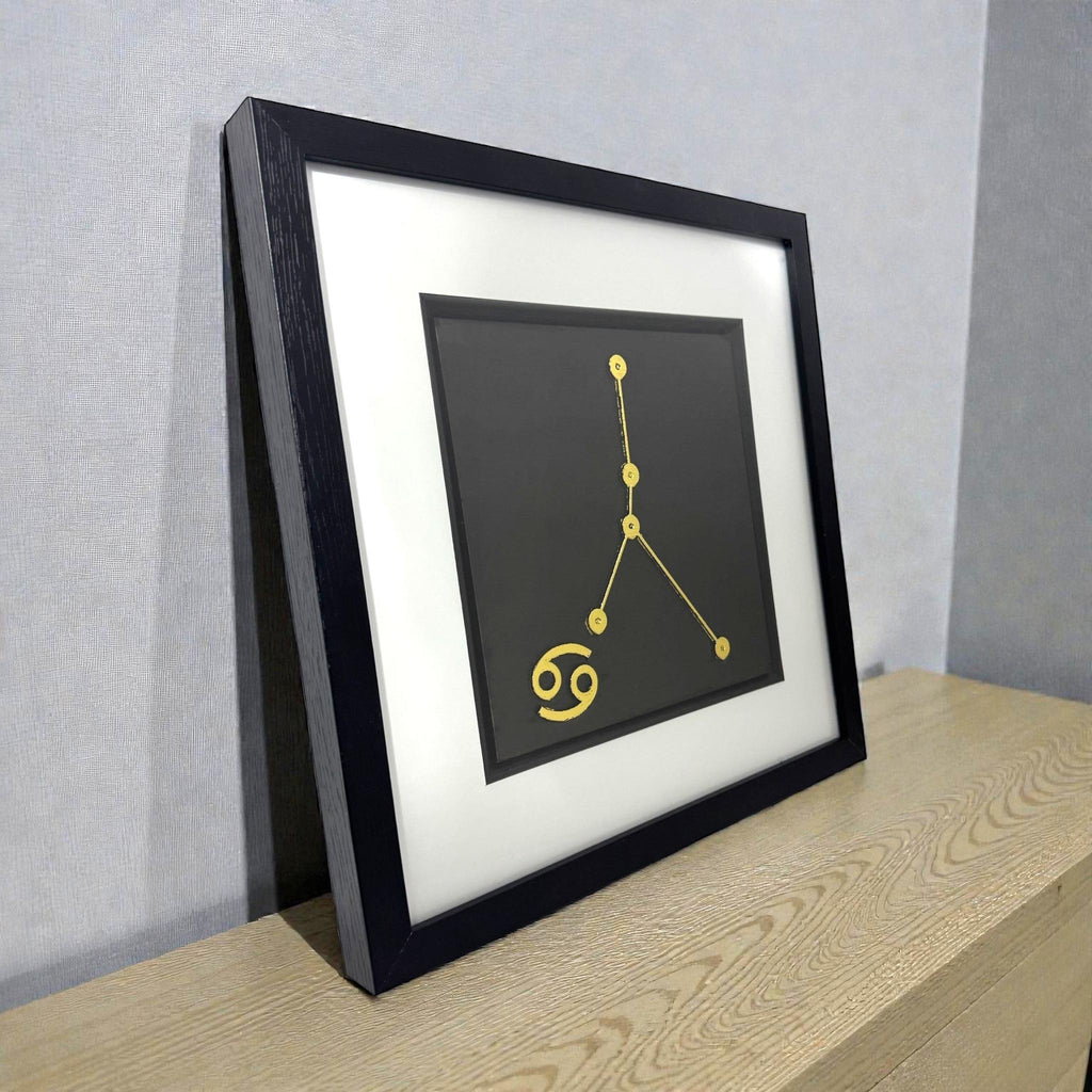 Framed Cancer constellation wall art in gold leaf with zodiac symbol on a black background, displayed on a shelf in a modern interior
