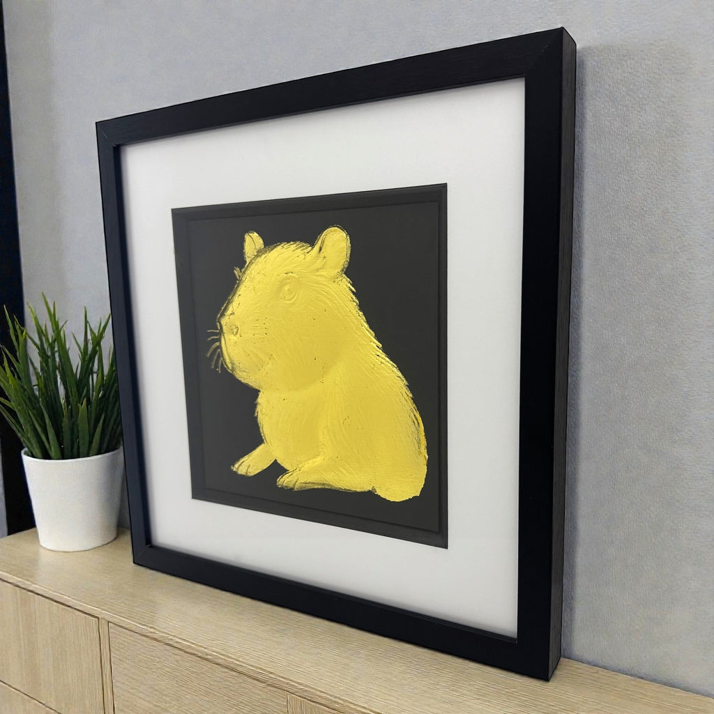 Capybara portrait wall art in gold leaf styled on a light wood cabinet beside a plant in a black frame with white mount