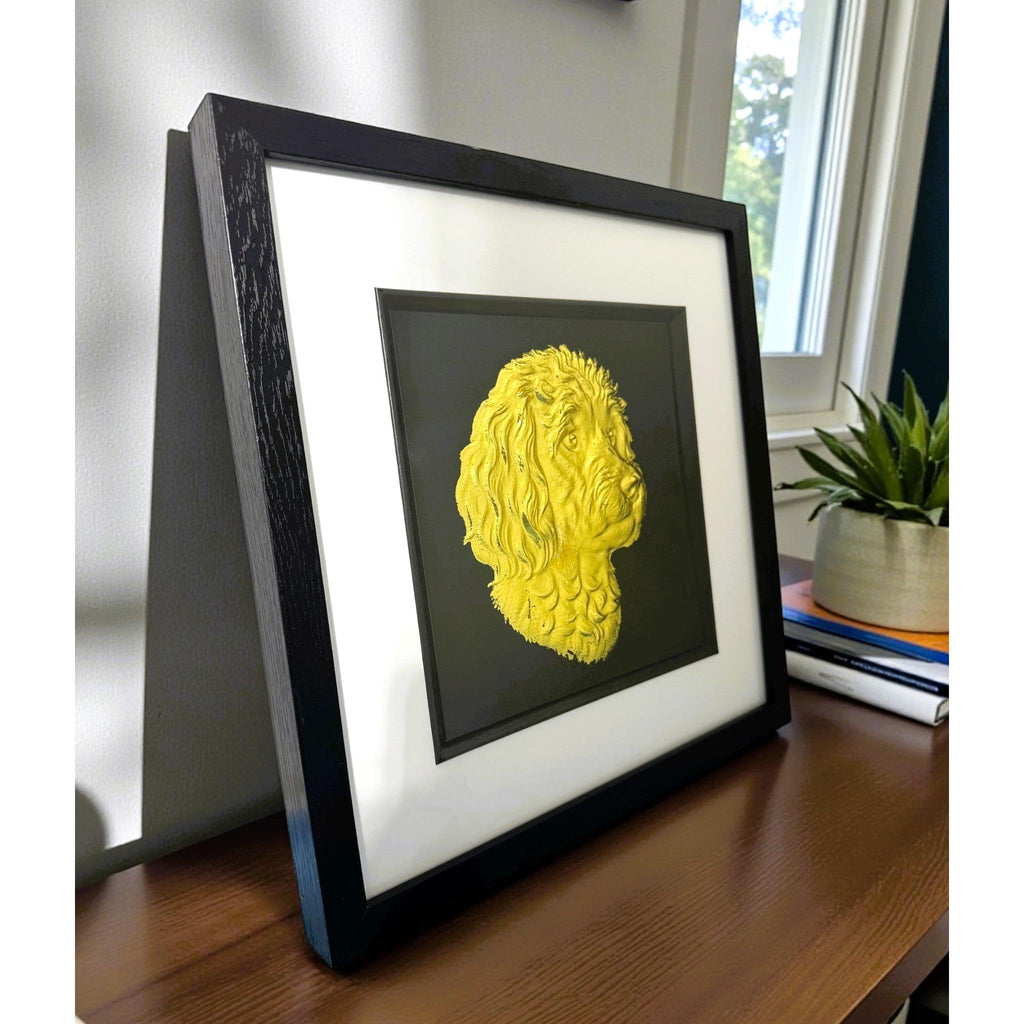 Gold cavapoo wall art displayed in a black frame with white mount, shown angled on a wooden surface near a window to show depth and scale