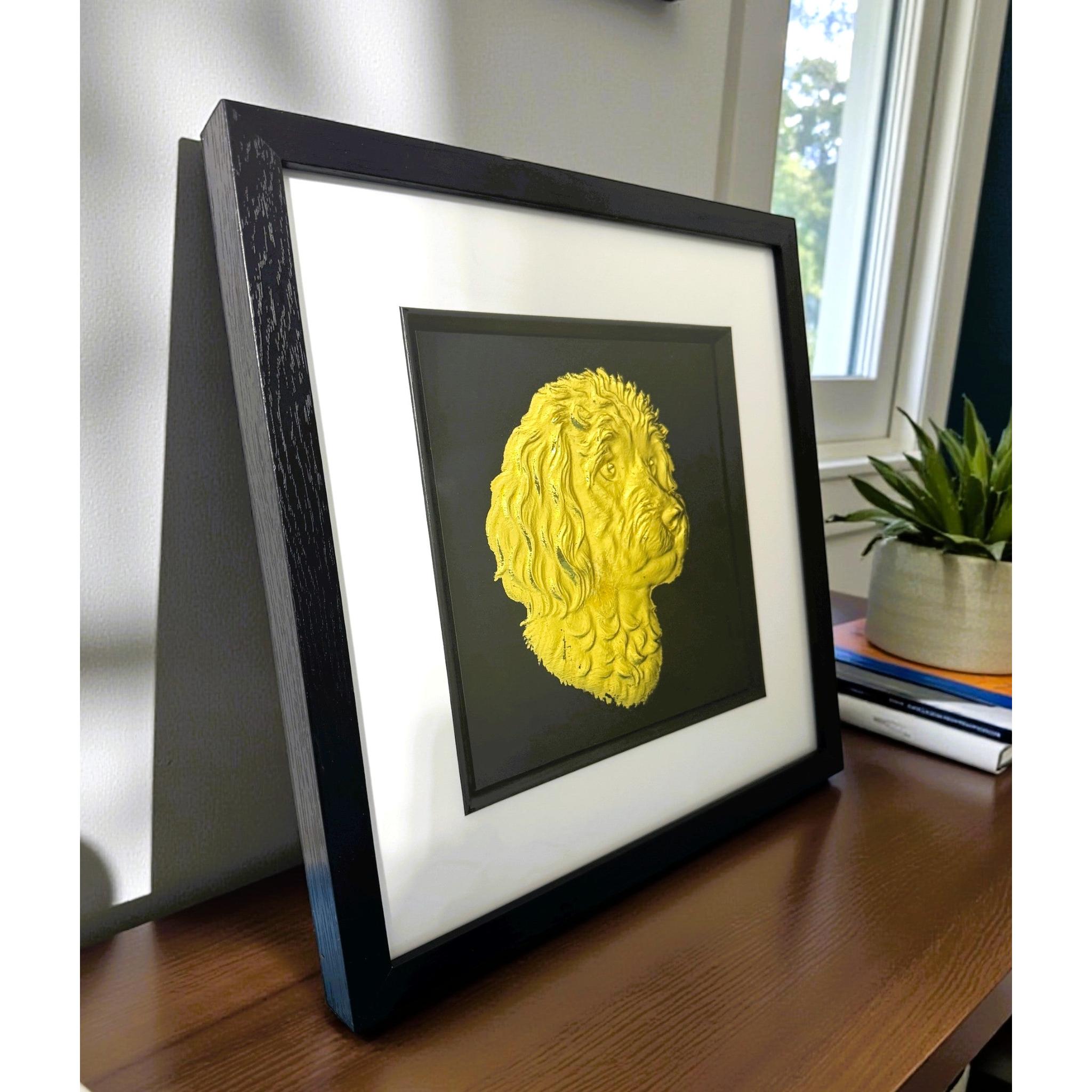 Gold cavapoo wall art displayed in a black frame with white mount, shown angled on a wooden surface near a window to show depth and scale