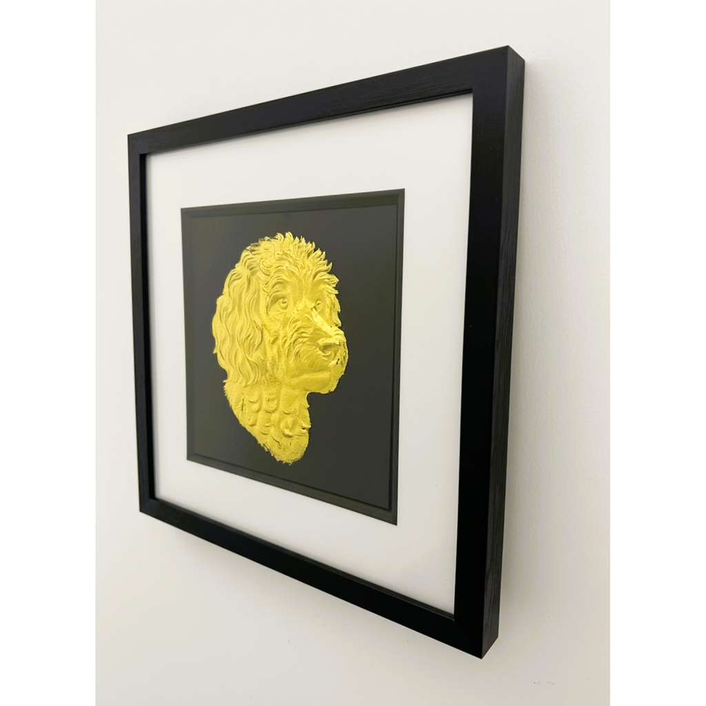 Framed cavapoo wall art featuring a gold relief portrait on a black background, shown hanging on a light wall in a black frame with white mount