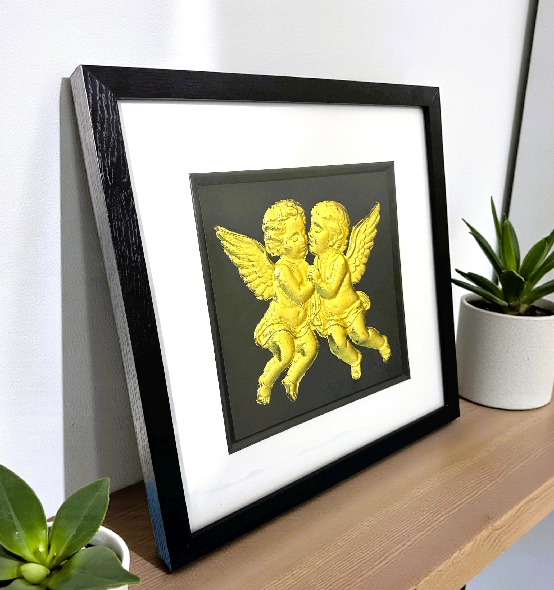 Angled desk view of gold leaf cherubs framed wall art displayed on a wooden shelf