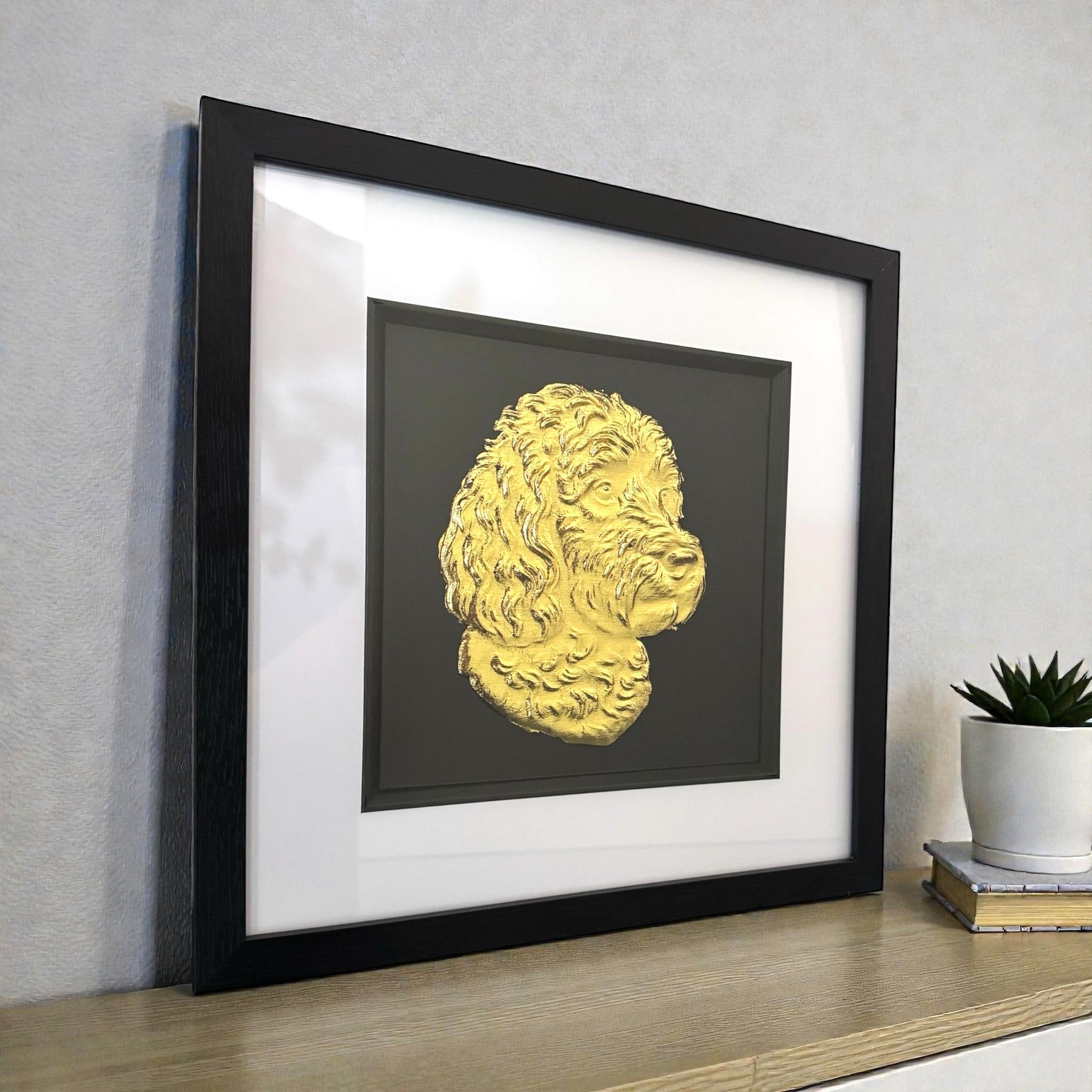Framed cockapoo wall art featuring a gold relief portrait on a black background, shown angled on a wooden surface with a small plant for scale