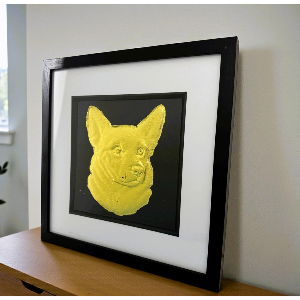 Gold corgi wall art displayed in a black frame with white mount, shown angled on a wooden surface against a light wall to show depth and scale