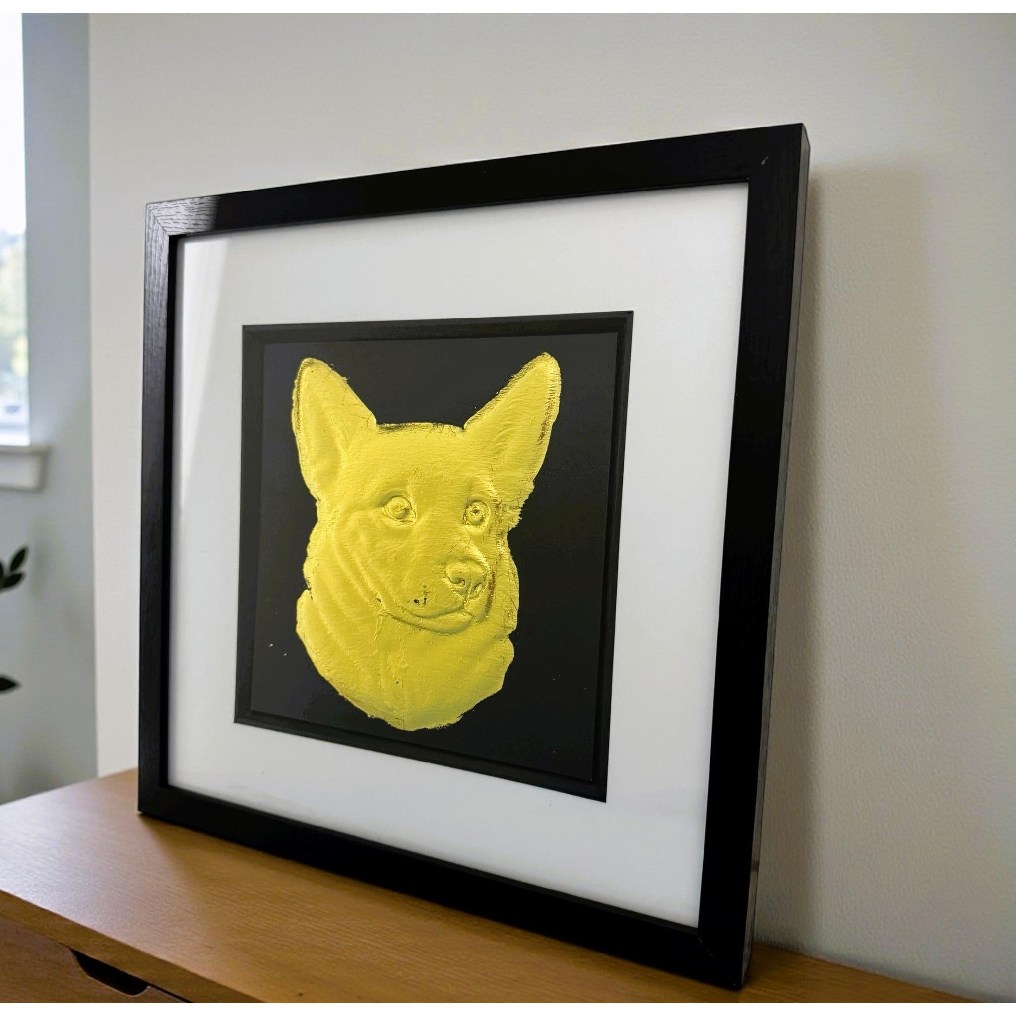 Gold corgi wall art displayed in a black frame with white mount, shown angled on a wooden surface against a light wall to show depth and scale