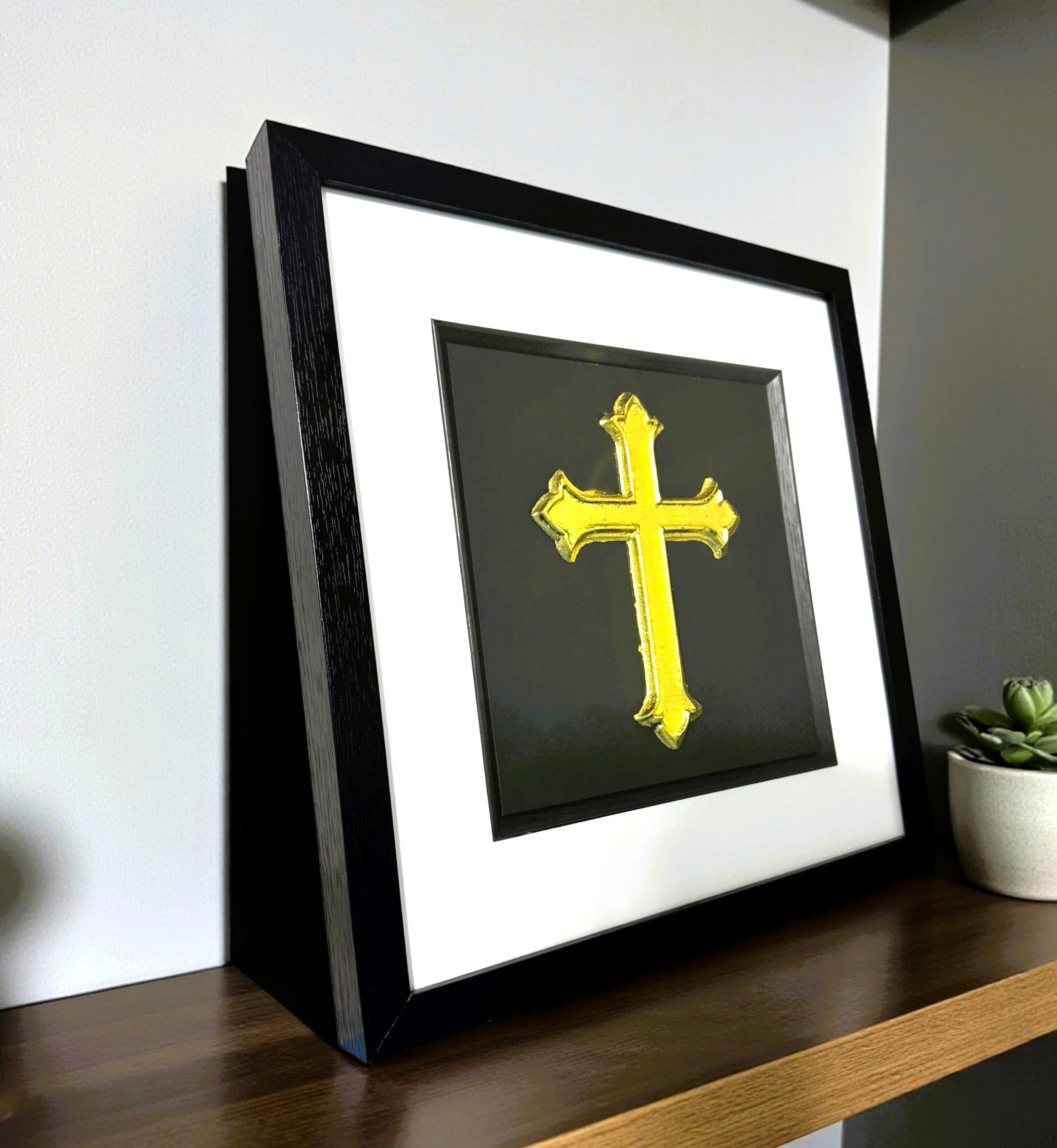 Gold leaf cross framed wall art photographed at an angle on a wooden shelf, black frame with white mount and black background