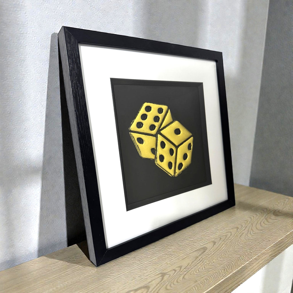 Framed dice wall art in gold leaf relief on a black background, displayed on a shelf in a modern interior