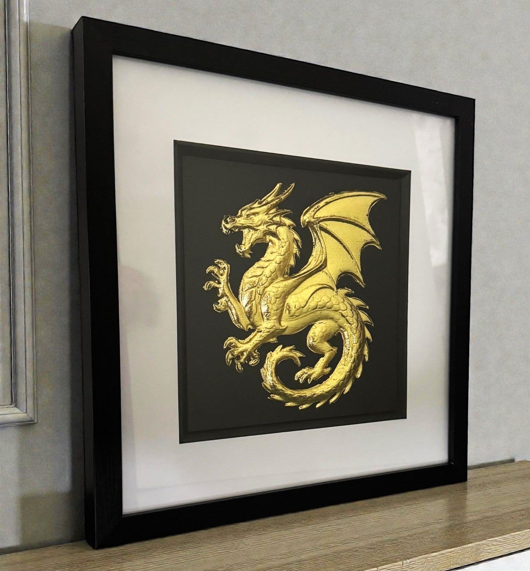 Framed dragon wall art in gold leaf relief on a black background, displayed on a shelf in a modern interior