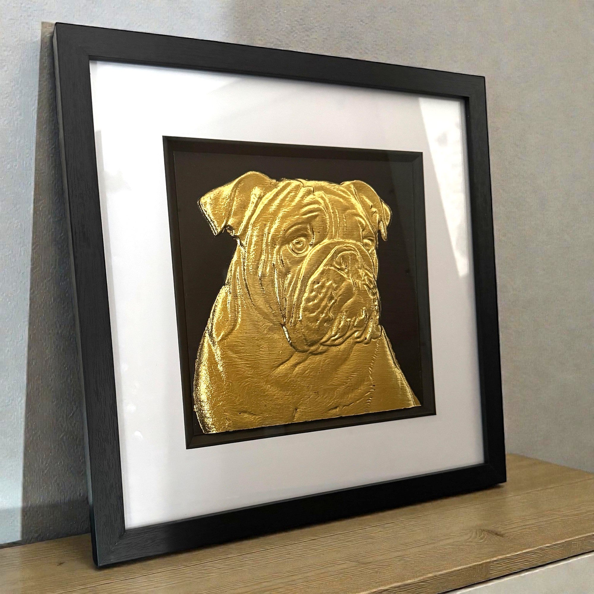 Framed English bulldog wall art in gold leaf relief on a black background with white mount and black frame, displayed on a shelf in a modern interior