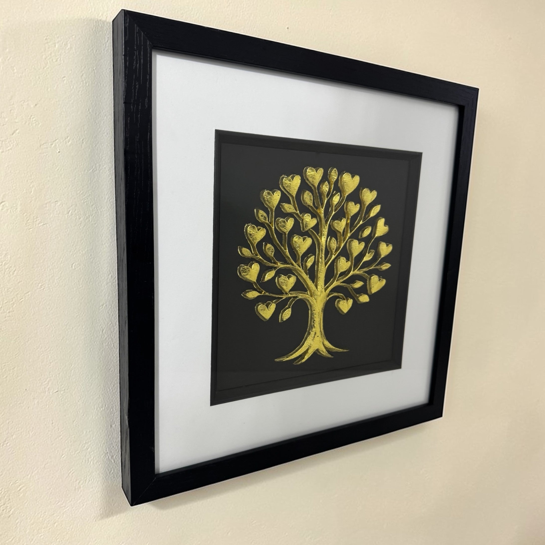 Angled view of a gold tree of life relief in a black box frame hanging on a neutral wall.