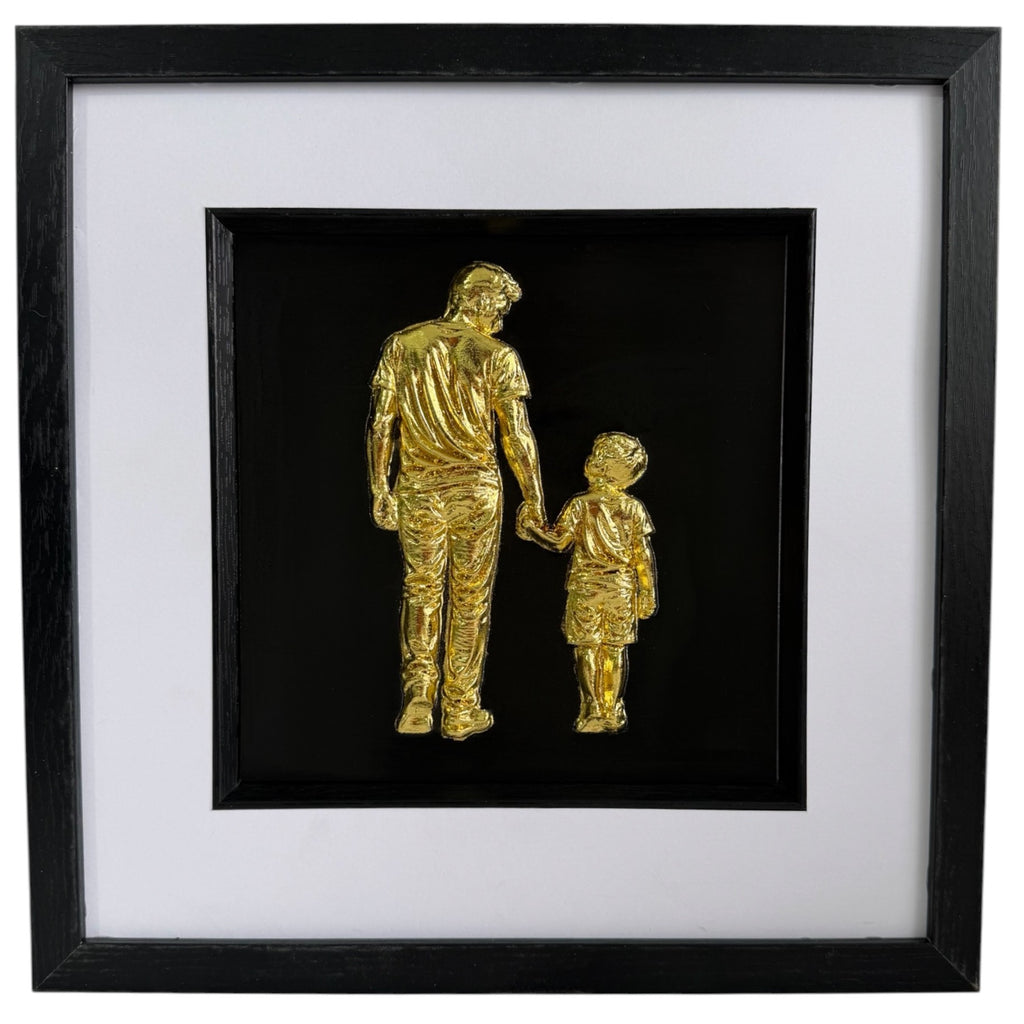 Gold father holding hands with son relief artwork in a black frame with white mount.