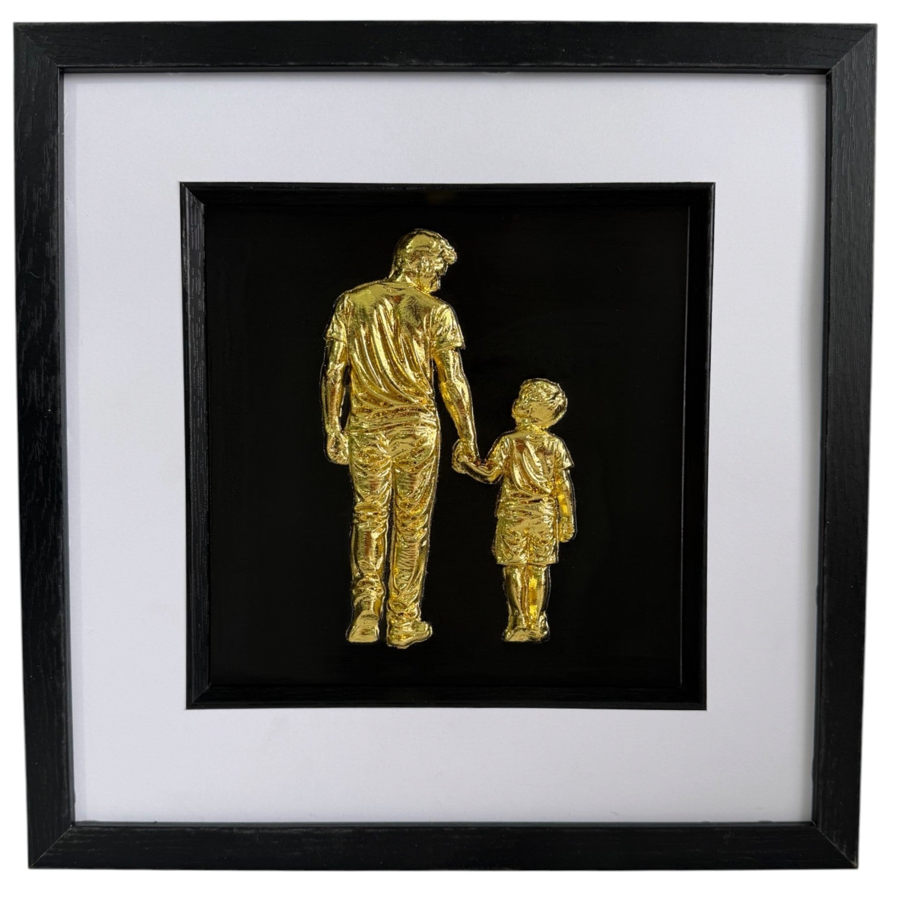 Gold father holding hands with son relief artwork in a black frame with white mount.
