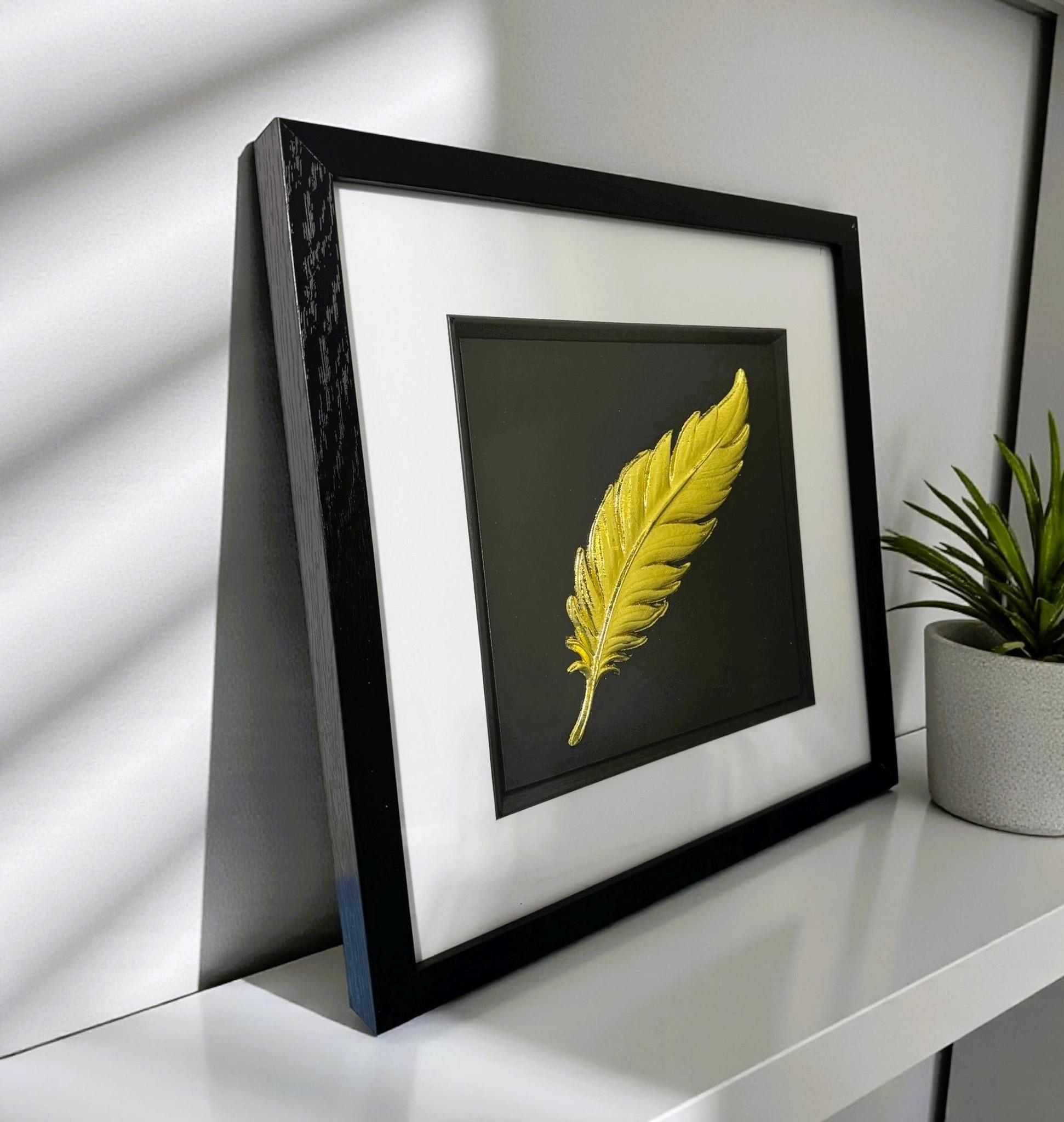 Floating feather wall art displayed on a desk in a black frame with gold leaf finish by JP Designs