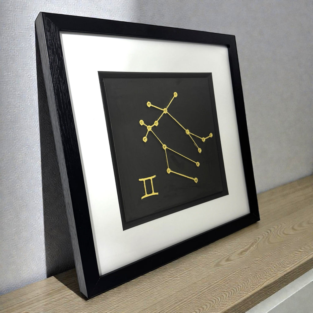 Framed Gemini constellation wall art in gold leaf with zodiac symbol on a black background, displayed on a shelf in a modern interior
