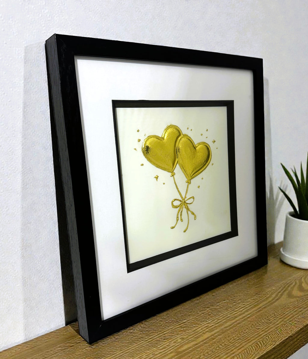Angled desk view of gold leaf heart balloons wall art on a light ivory background in a black wooden frame