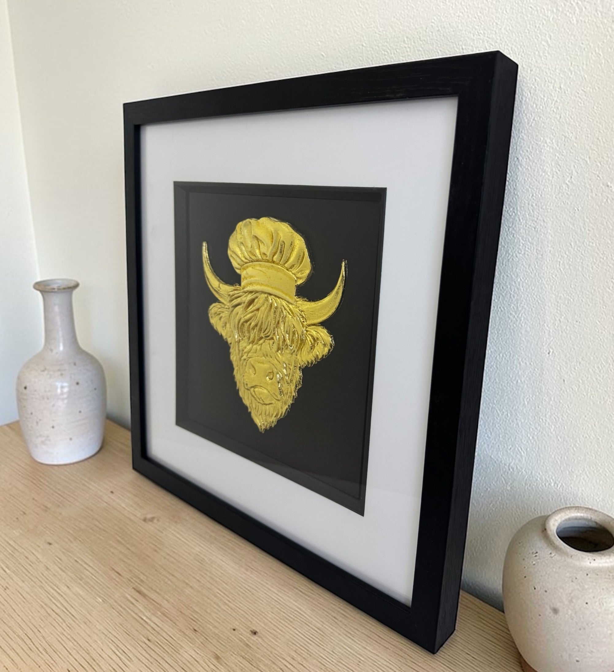 Gold Highland cow wall decor styled in a black box frame on a wooden sideboard between two ceramic vases.