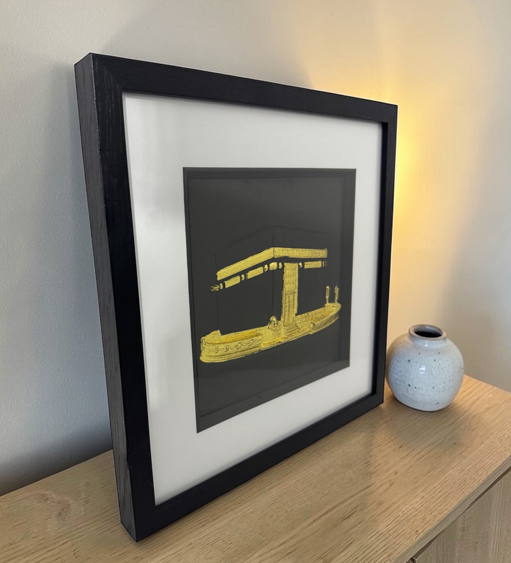 Angled view of a gold Kaaba relief in a black box frame with glass front on a wooden sideboard beside a vase and warm lamp.