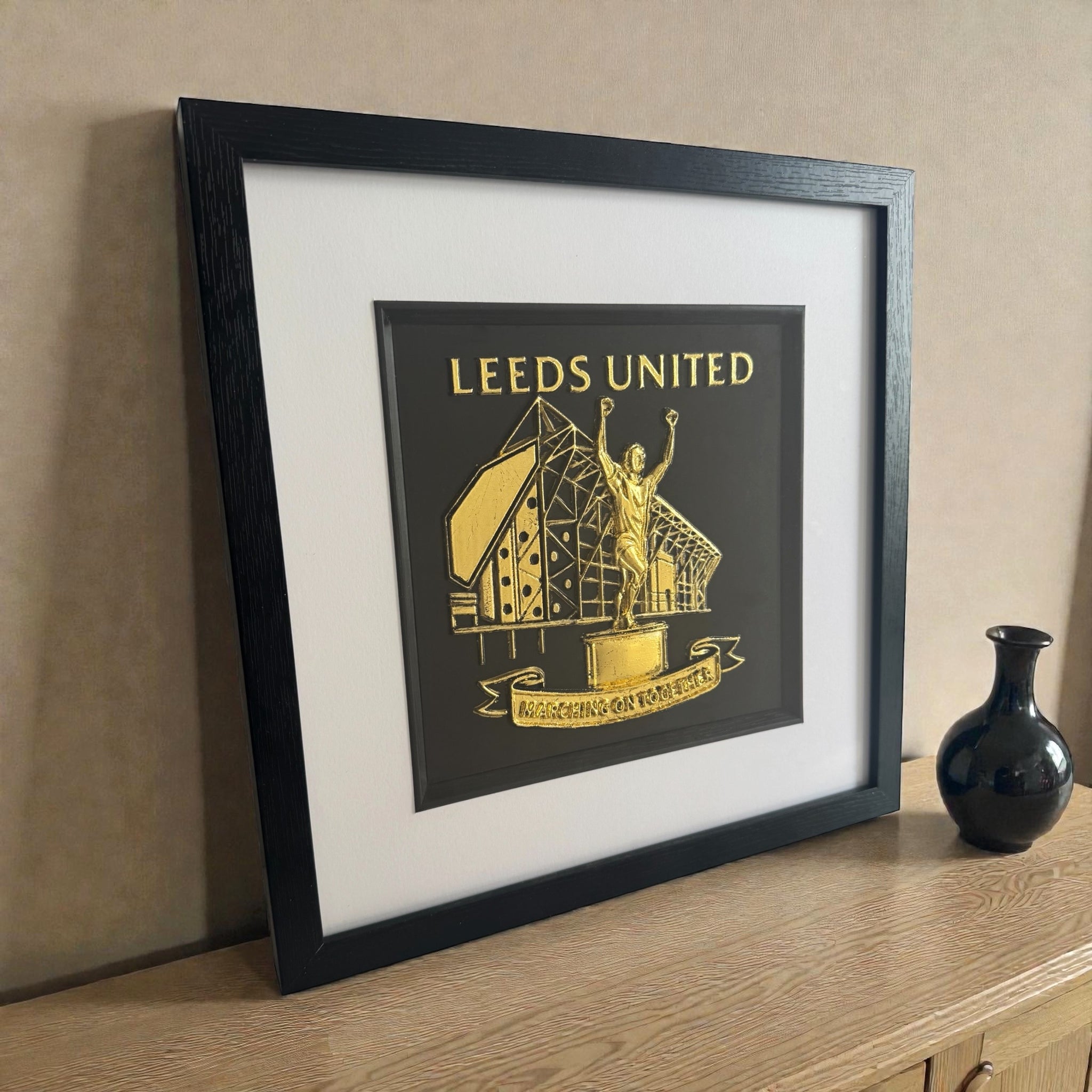 Framed Leeds United Gold Leaf relief artwork styled on a wooden sideboard, black box frame with white mount and inner black surround