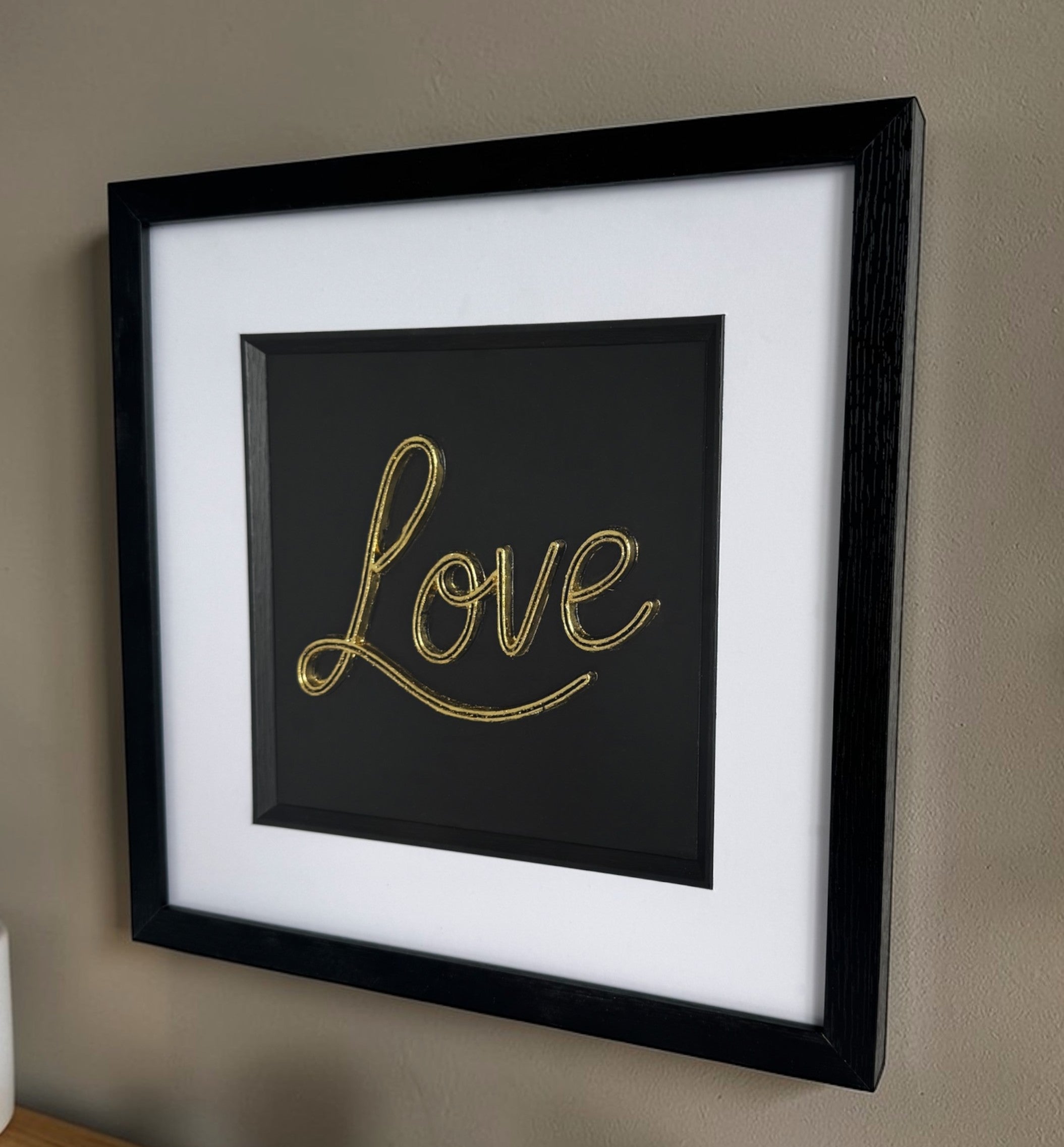 Angled wall display of framed 3D Love sign in gold finish on a beige wall with soft natural light.