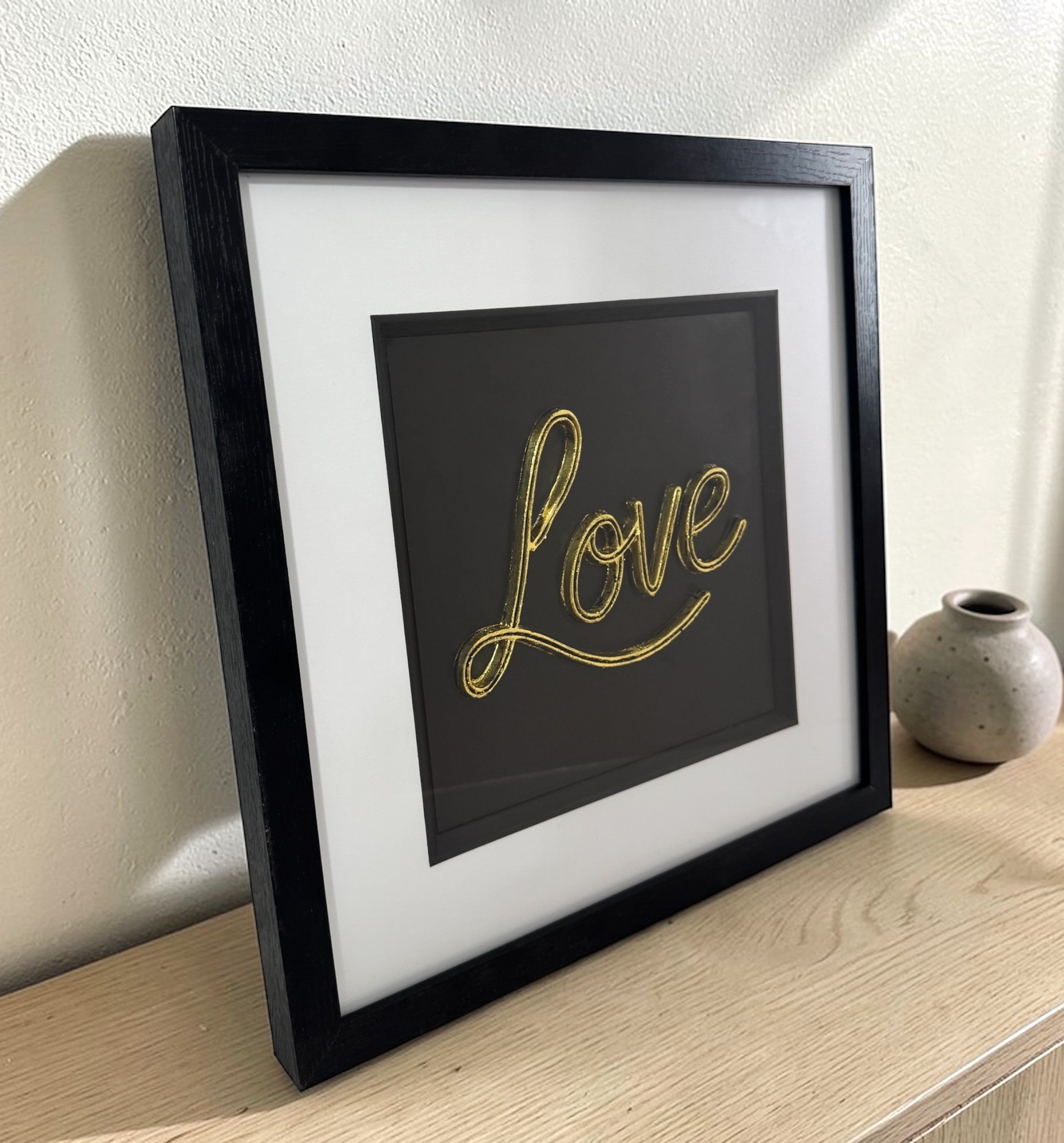 Angled view of framed 3D Love sign in gold finish on a wooden sideboard beside a ceramic vase.